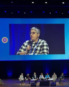 Paneelikeskustelu konferenssin päälavalla ja näytöllä suurennettuna puhuja.